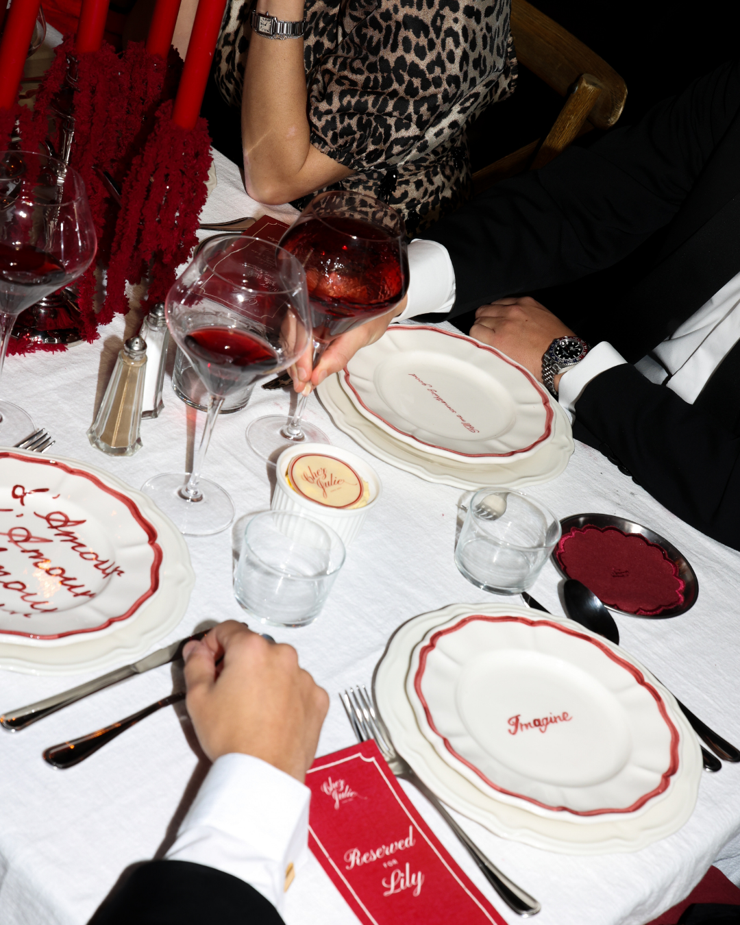 Chez Jules tablescape close-up – scalloped plates with red detailing, wine glasses and deep red accessories by Imaginary Friends.
