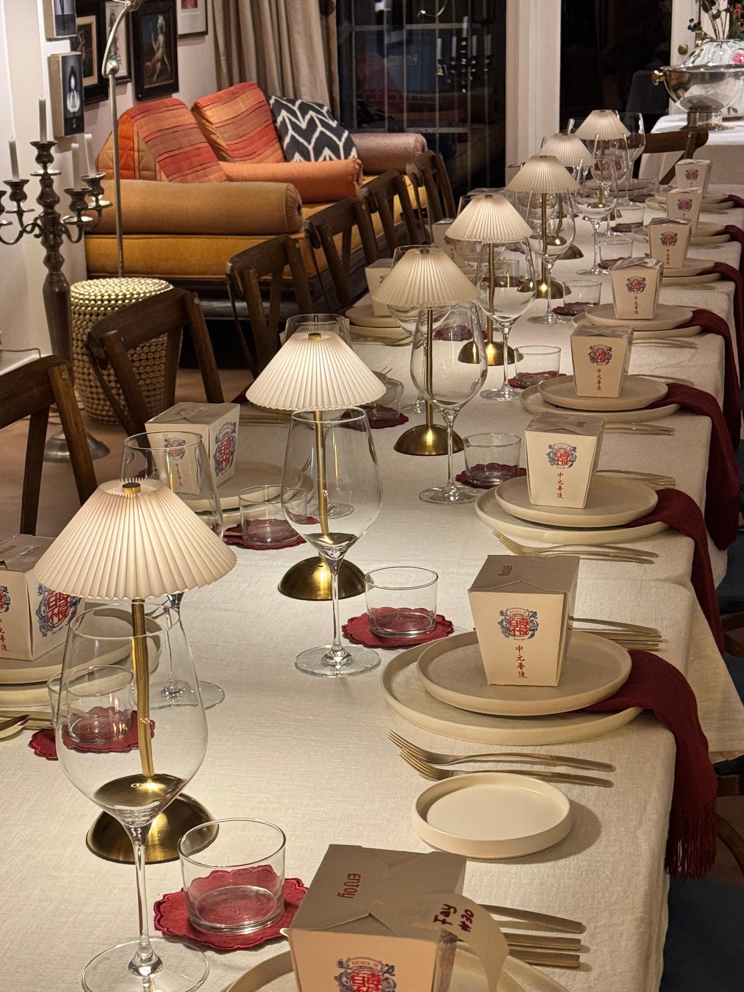 Elegant indoor dinner table styling by Imaginary Friends featuring brass table lamps, stacked neutral plates, red linen details, glassware, and personalized takeaway boxes.