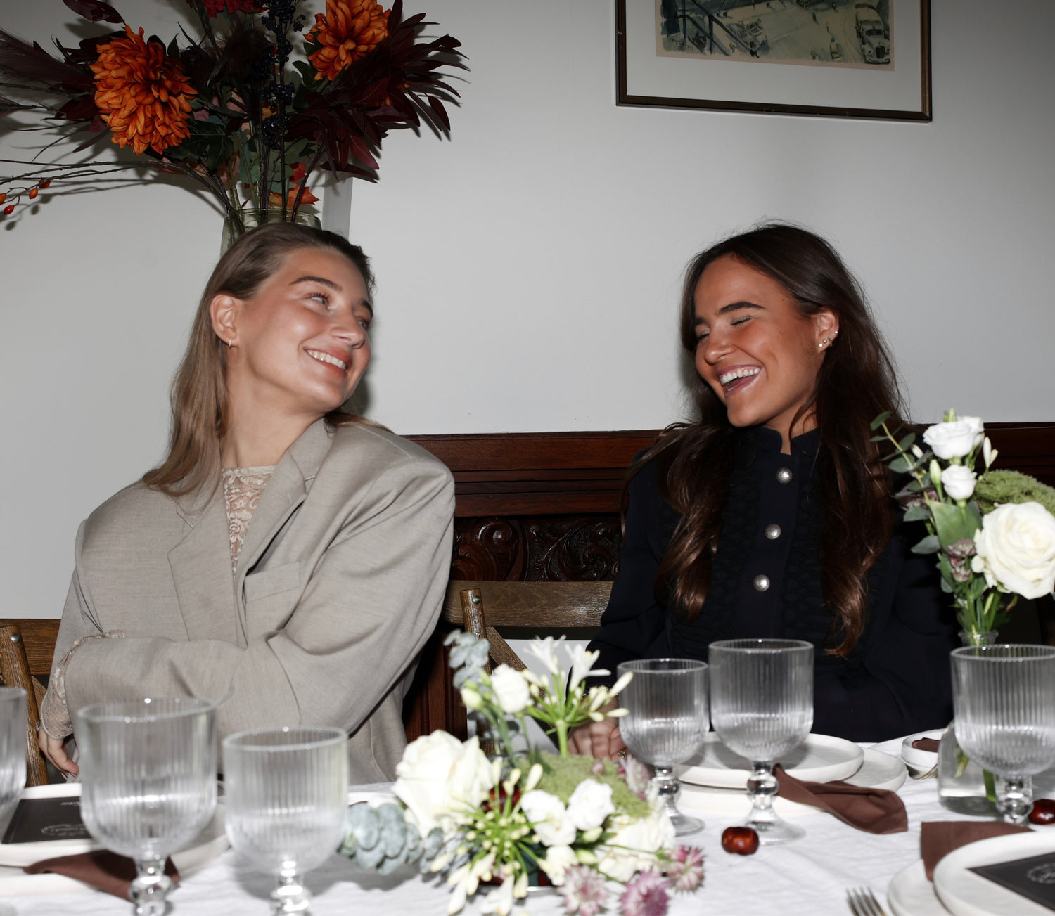 Pien de Pont and Bo Boender, founders of Imaginary Friends, laughing together at an elegantly styled dinner table with floral arrangements and glassware.